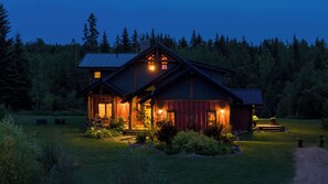 A two-story log cabin with a porch and a well-lit interior, surrounded by a forested landscape. - Luxury West Coast Post & Beam Estate on 142 Acres of Private Alberta Wilderness! (Westlock County)