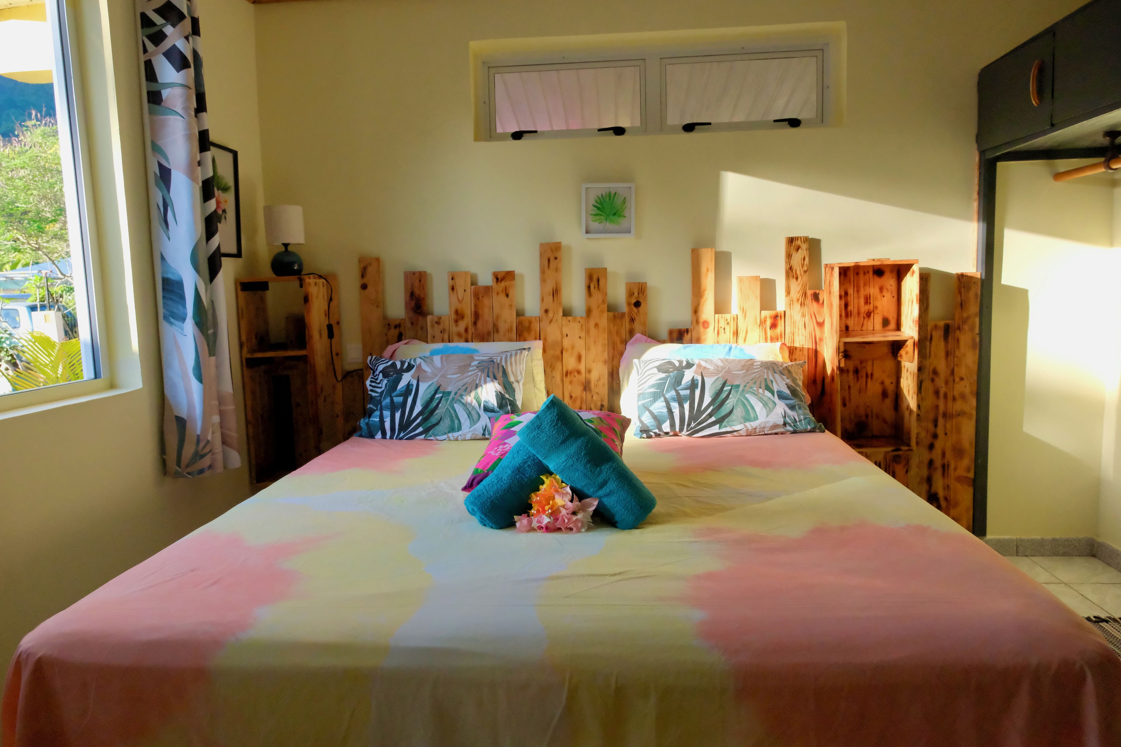 A bed with a colorful bedspread, a blue towel, and decorative pillows. There is a window with a view of greenery outside.