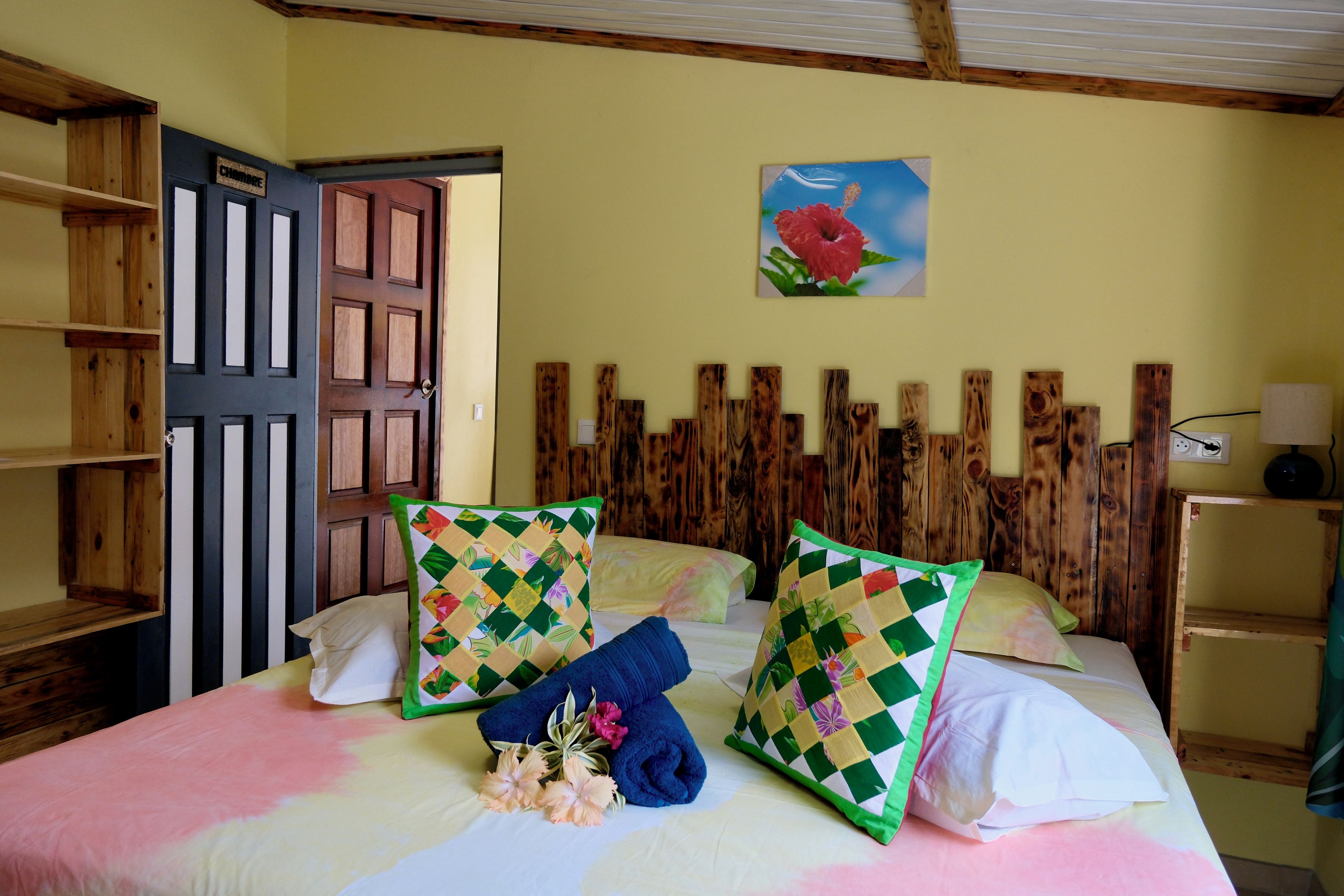 A room with a bed, wooden headboard, a painting of a flower, and a shelf with a lamp.