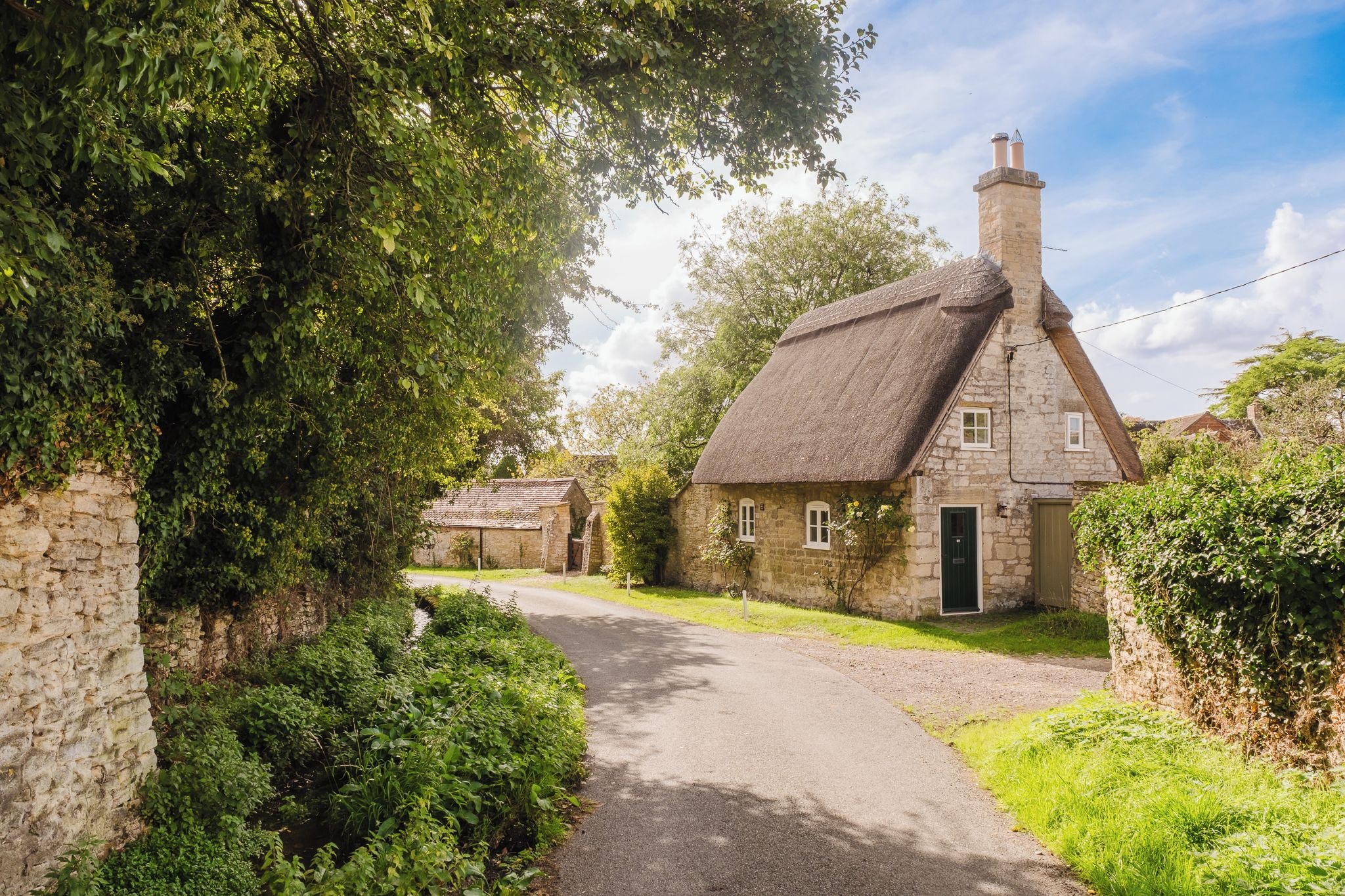 A rural stone house with a thatched roof, surrounded by greenery and a narrow road.