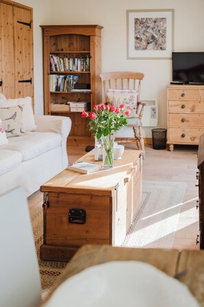 A living room with a white sofa, a wooden coffee table with a vase of flowers, a bookshelf, a wooden cabinet, a chair, and a television. - Hoglets - Cosy Countryside Retreat (Conderton)