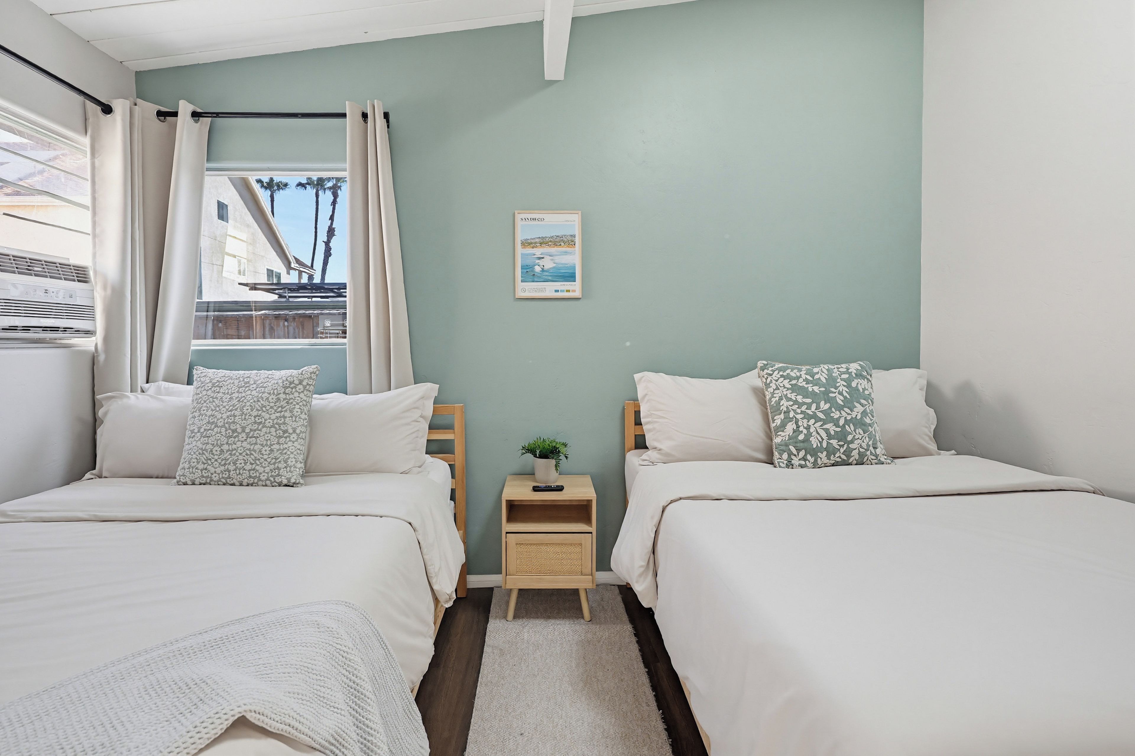A bedroom with two beds, a small bedside table with a plant, a framed picture on the wall, and a window with curtains.