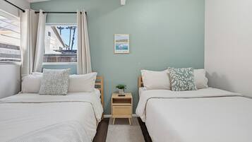 A bedroom with two beds, a small bedside table with a plant, a framed picture on the wall, and a window with curtains.