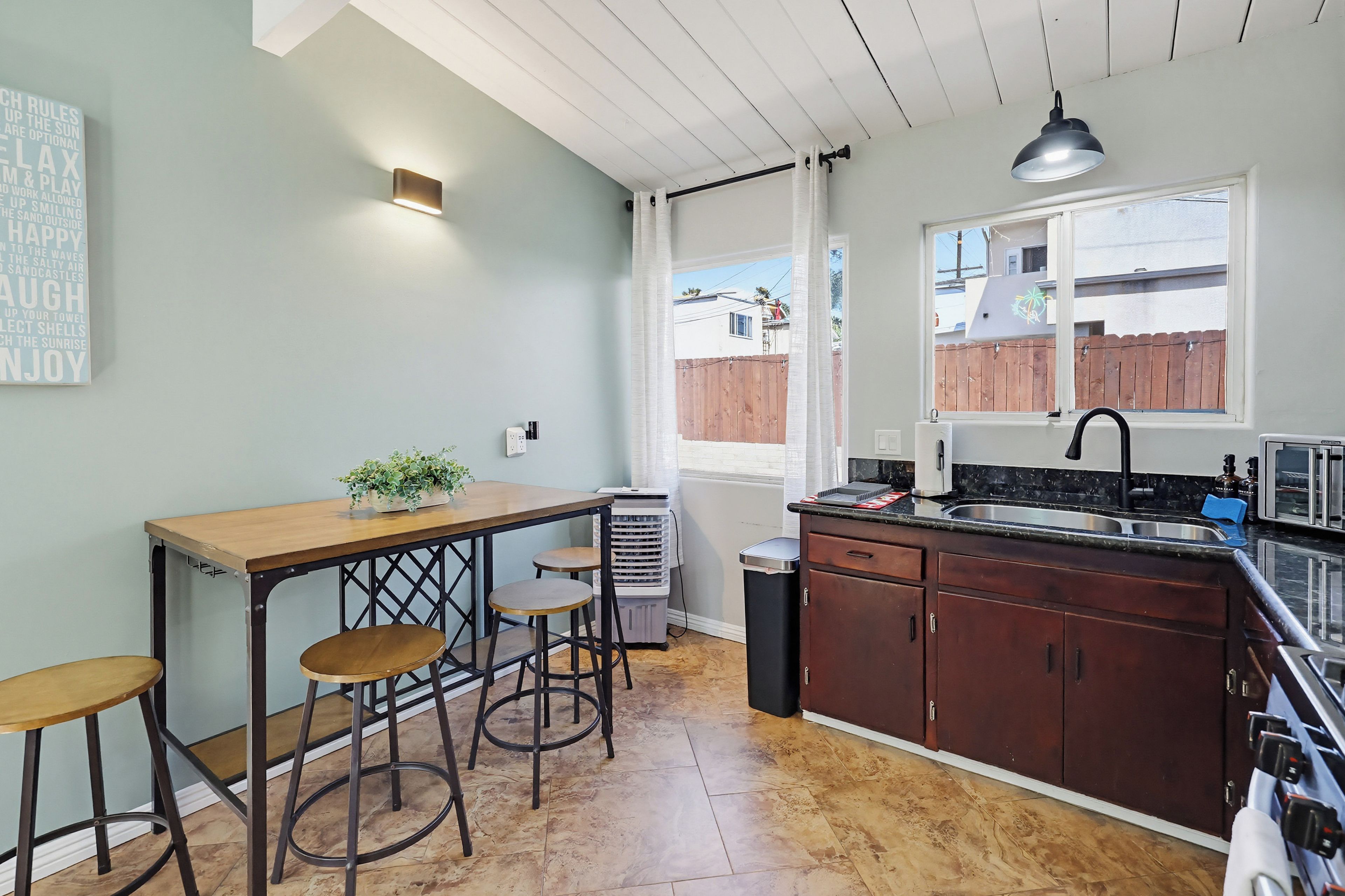 A kitchen with a wooden table, bar stools, a sink, and a window with a view of a wooden fence.
