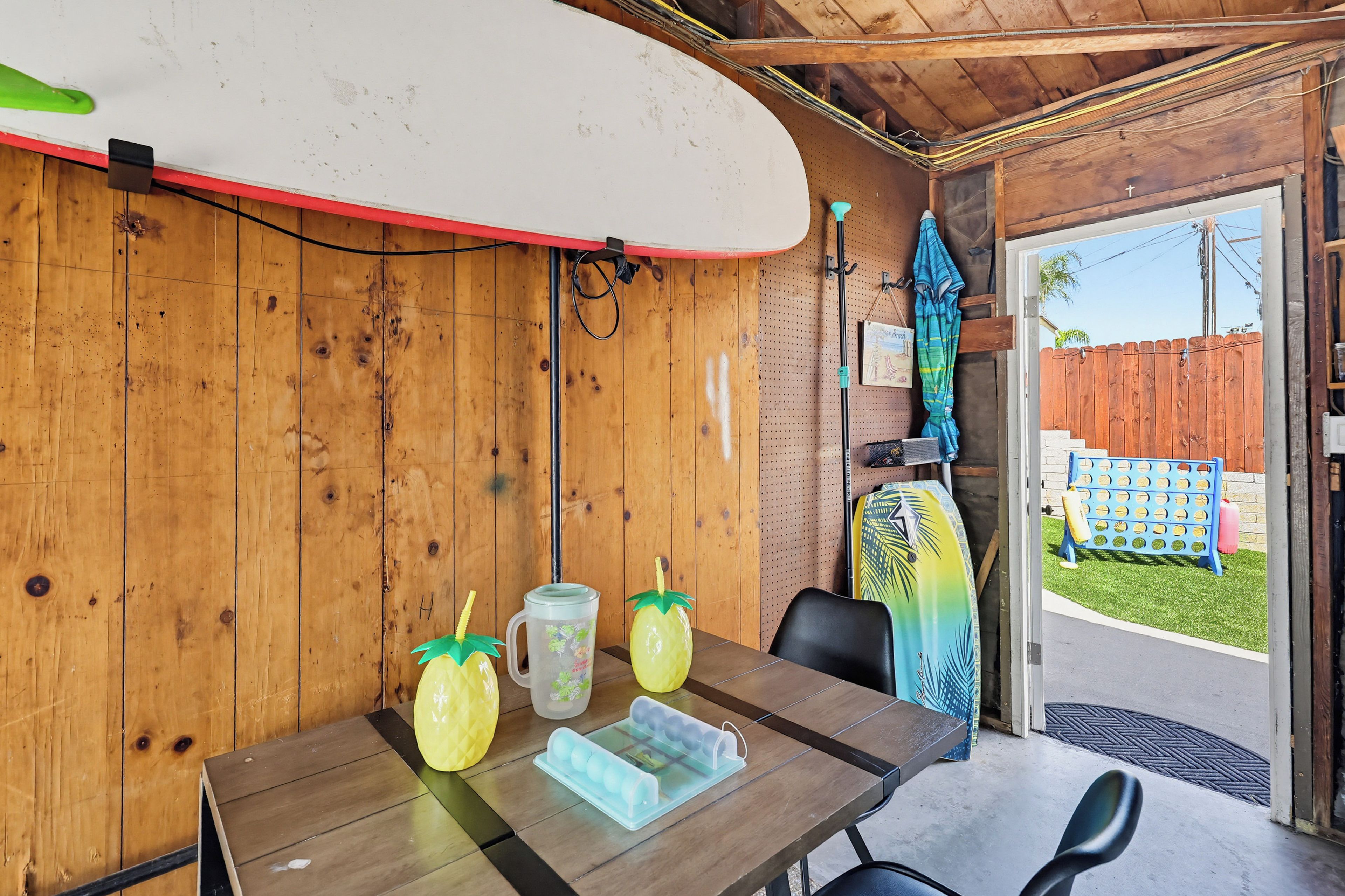 A wooden interior with a surfboard mounted on the wall, a table with a pitcher and glasses, and a door leading outside.