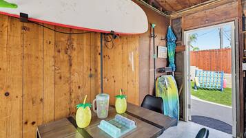 A wooden interior with a surfboard mounted on the wall, a table with a pitcher and glasses, and a door leading outside.