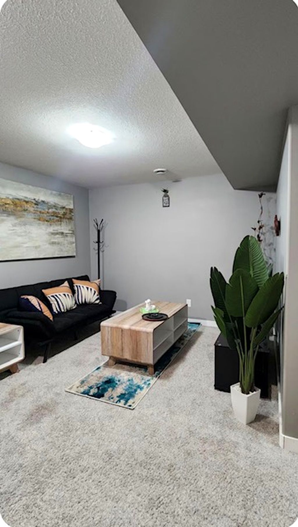 A living room with a black sofa, a wooden coffee table, a large green plant, and a painting on the wall.