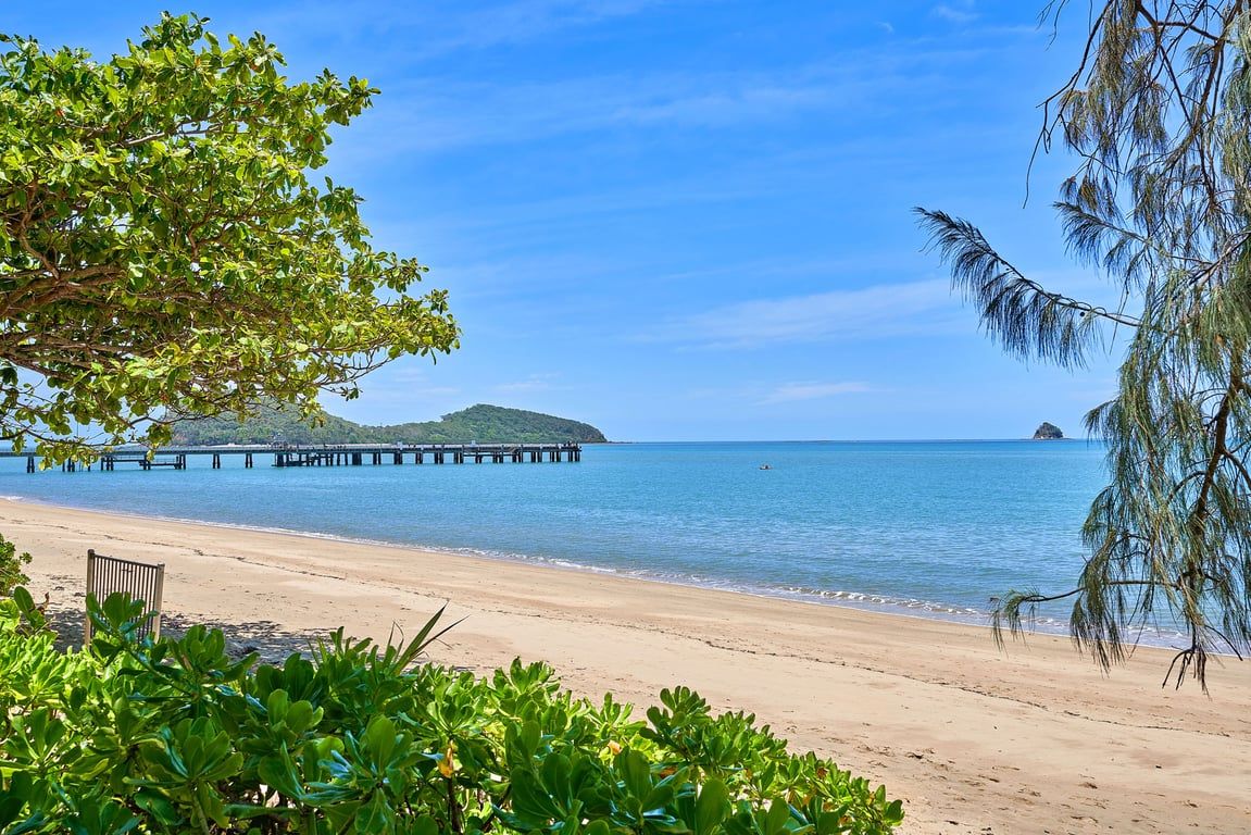 A beach with a wooden pier extending into calm waters, surrounded by lush greenery.