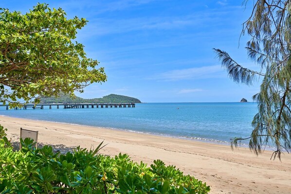 A beach with a wooden pier extending into calm waters, surrounded by lush greenery.
