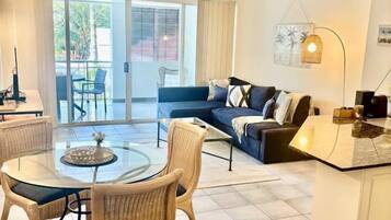 A living room with a glass-top table, wicker chairs, a sofa, and a kitchen area with a countertop.