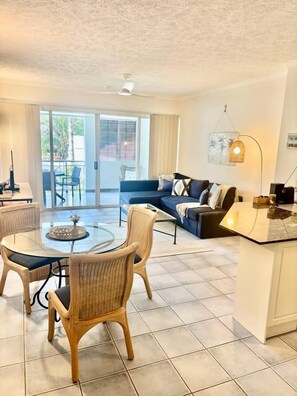 A living room with a glass-top table, wicker chairs, a sofa, and a kitchen area with a countertop.