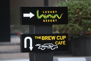 Dos senyals direccionals, un per 'Luxury Resort' i un altre per 'The Brew Cup Café', amb un símbol de 'Drive-Thru'.