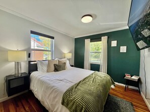 A bedroom with a bed, two bedside tables with lamps, a window with curtains, and a wall-mounted TV. - Rose Park Cottage | Near Downtown | Mid-Term (Salt Lake City)