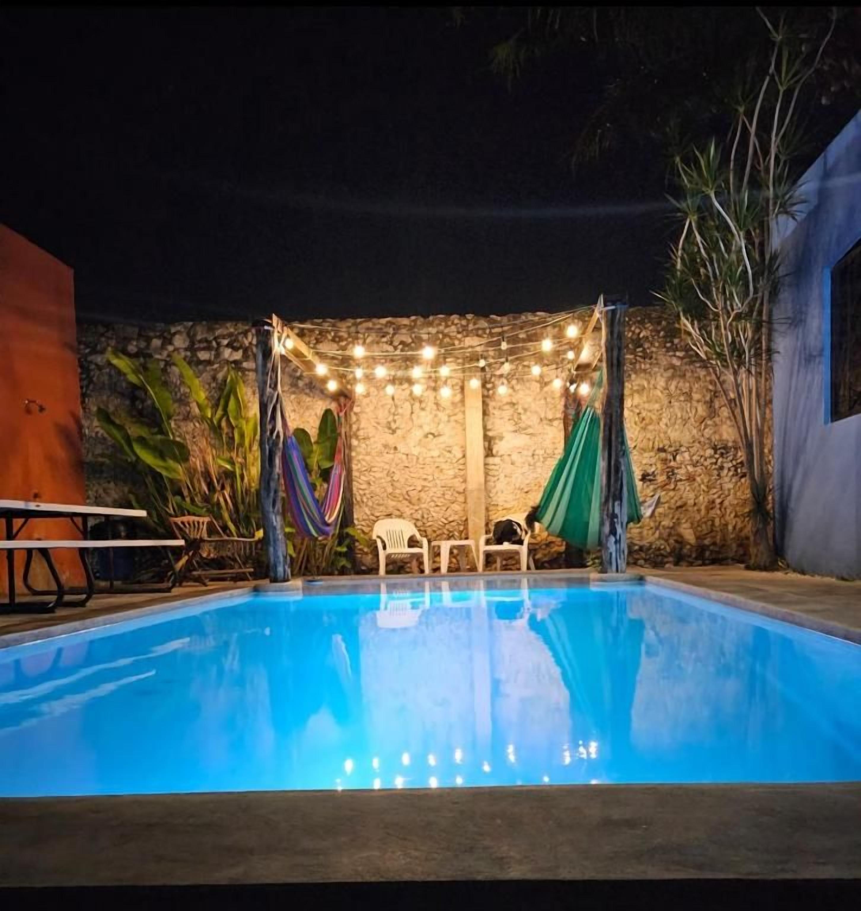 Una piscina con un muro de piedra de fondo, luces colgantes y una hamaca.