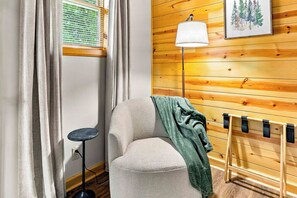 A cozy room with a wooden wall, a window with blinds, a lamp, a chair, and a small table. - Timber Nook -2BR Near Sand Valley & Lake Arrowhead (Nekoosa)