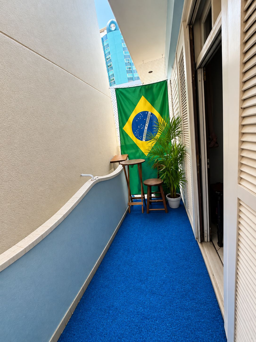 A narrow balcony with a blue carpet, a small table, a potted plant, and a Brazilian flag.