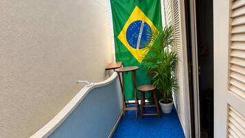 A narrow balcony with a blue carpet, a small table, a potted plant, and a Brazilian flag.