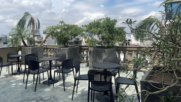 Uma área externa com mesas e cadeiras redondas, cercada por plantas em vasos e árvores.