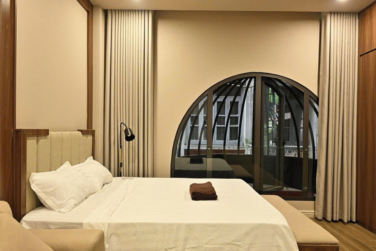 Quarto de hotel com cama, mesa de cabeceira, luminária e janela com vista para o exterior.