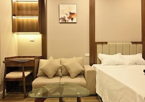 A hotel room with a sofa, a glass coffee table, a bed with white bedding, and a painting on the wall.
