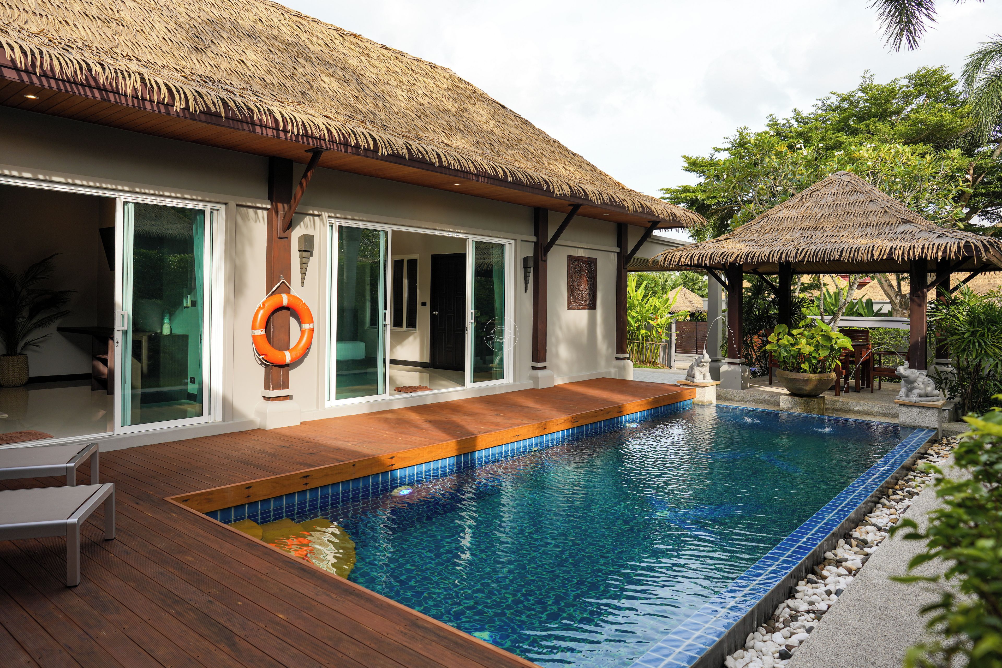 Un espace aménagé au bord de la piscine, avec une terrasse en bois, une structure dotée d’un toit de chaume et une bouée de sauvetage.