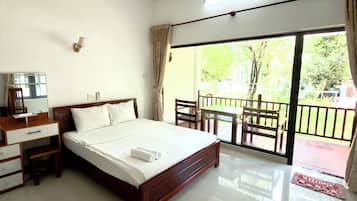 Habitación de hotel con cama, escritorio, silla, mesita pequeña y balcón con vistas a la vegetación.