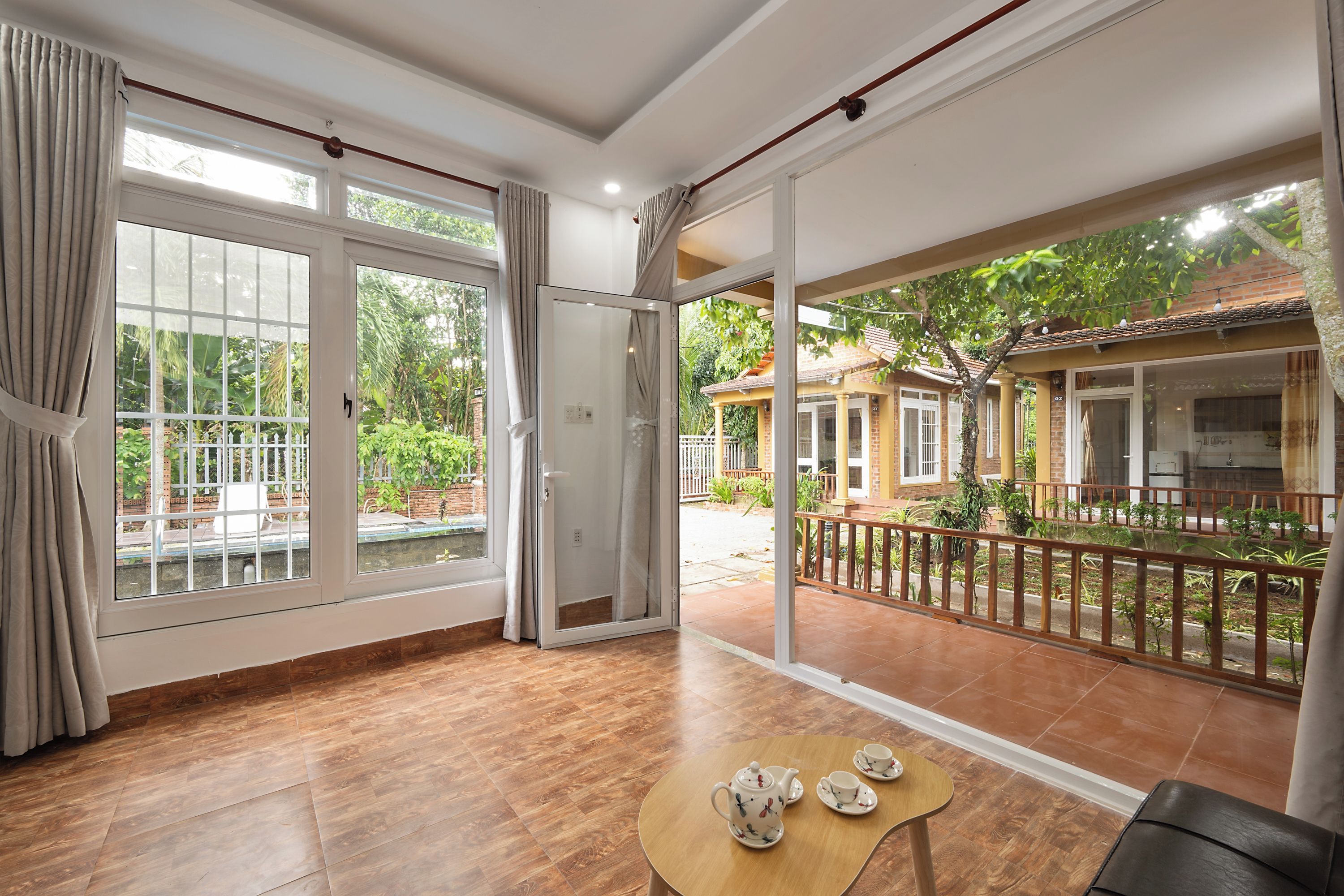 A spacious room with a large sliding glass door leading to a balcony, a wooden table with a teapot and cups, and a view of a garden and buildings outside.