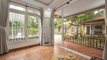 A spacious room with a large sliding glass door leading to a balcony, a wooden table with a teapot and cups, and a view of a garden and buildings outside.