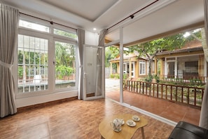 A spacious room with a large sliding glass door leading to a balcony, a wooden table with a teapot and cups, and a view of a garden and buildings outside.