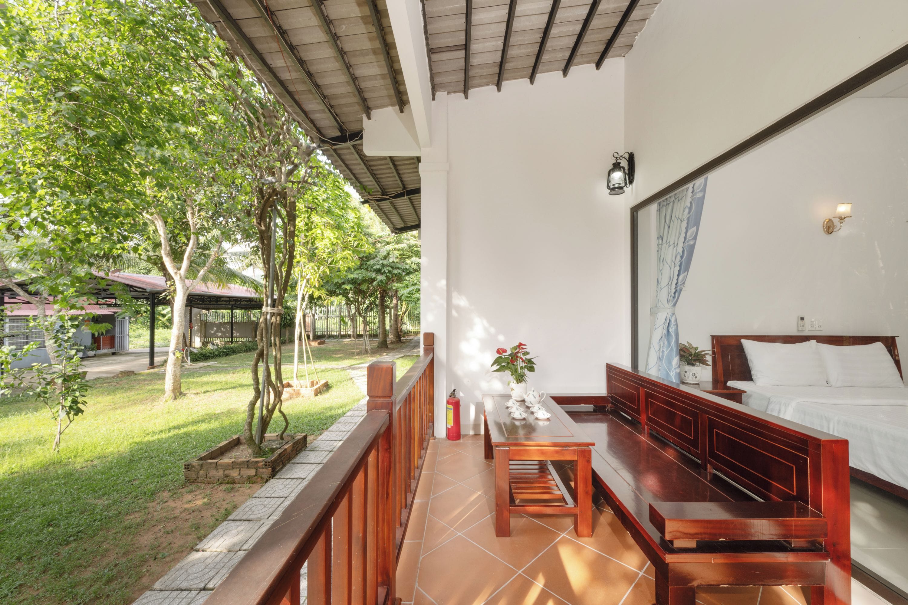 A room with a bed, a wooden bench, a table with teapots, and a view of a garden.