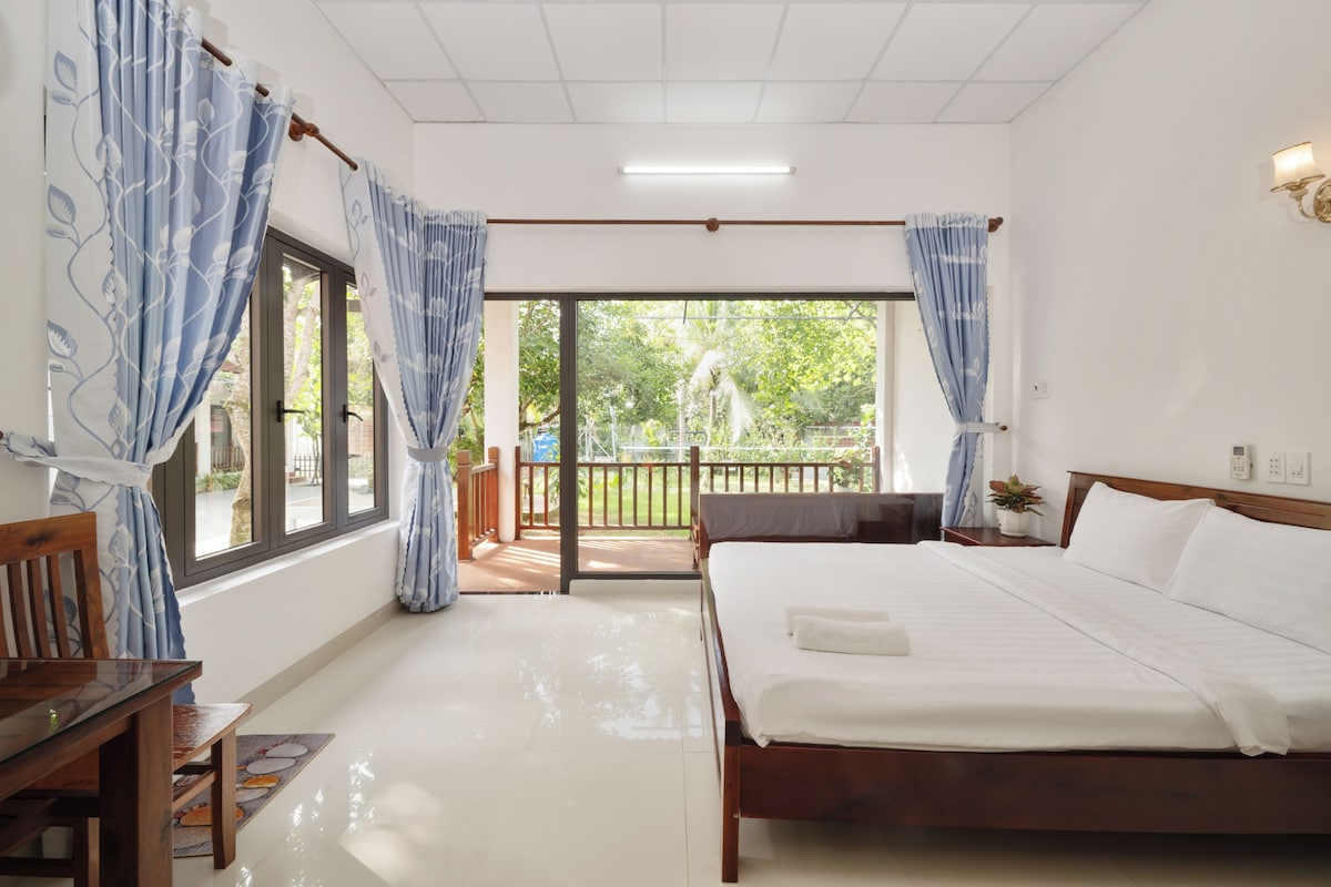 A hotel room with a bed, a desk, a chair, a balcony with a view of greenery, and a ceiling light.