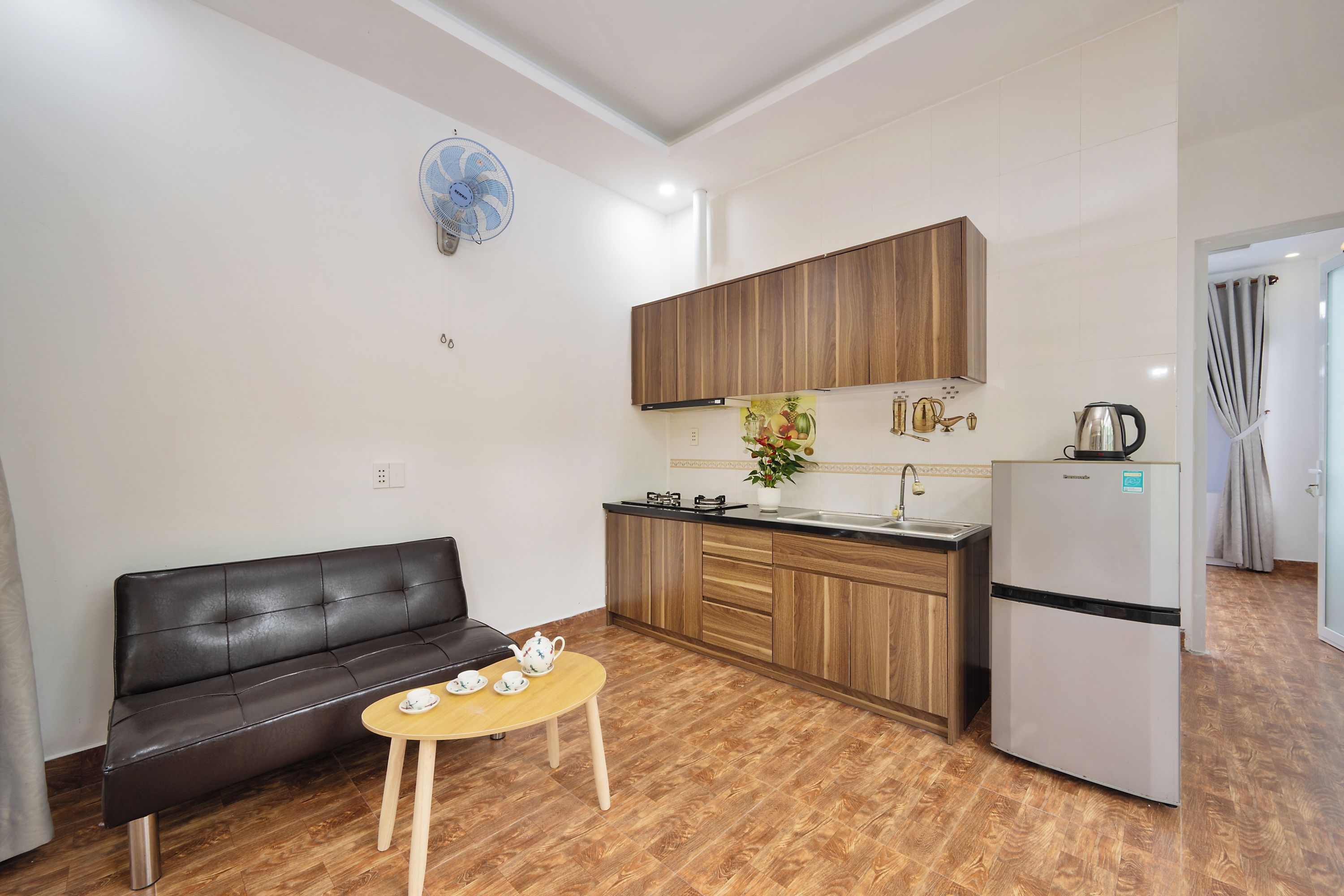 A compact living space with a kitchenette, a black leather sofa, a small round table with tea cups, and a refrigerator.