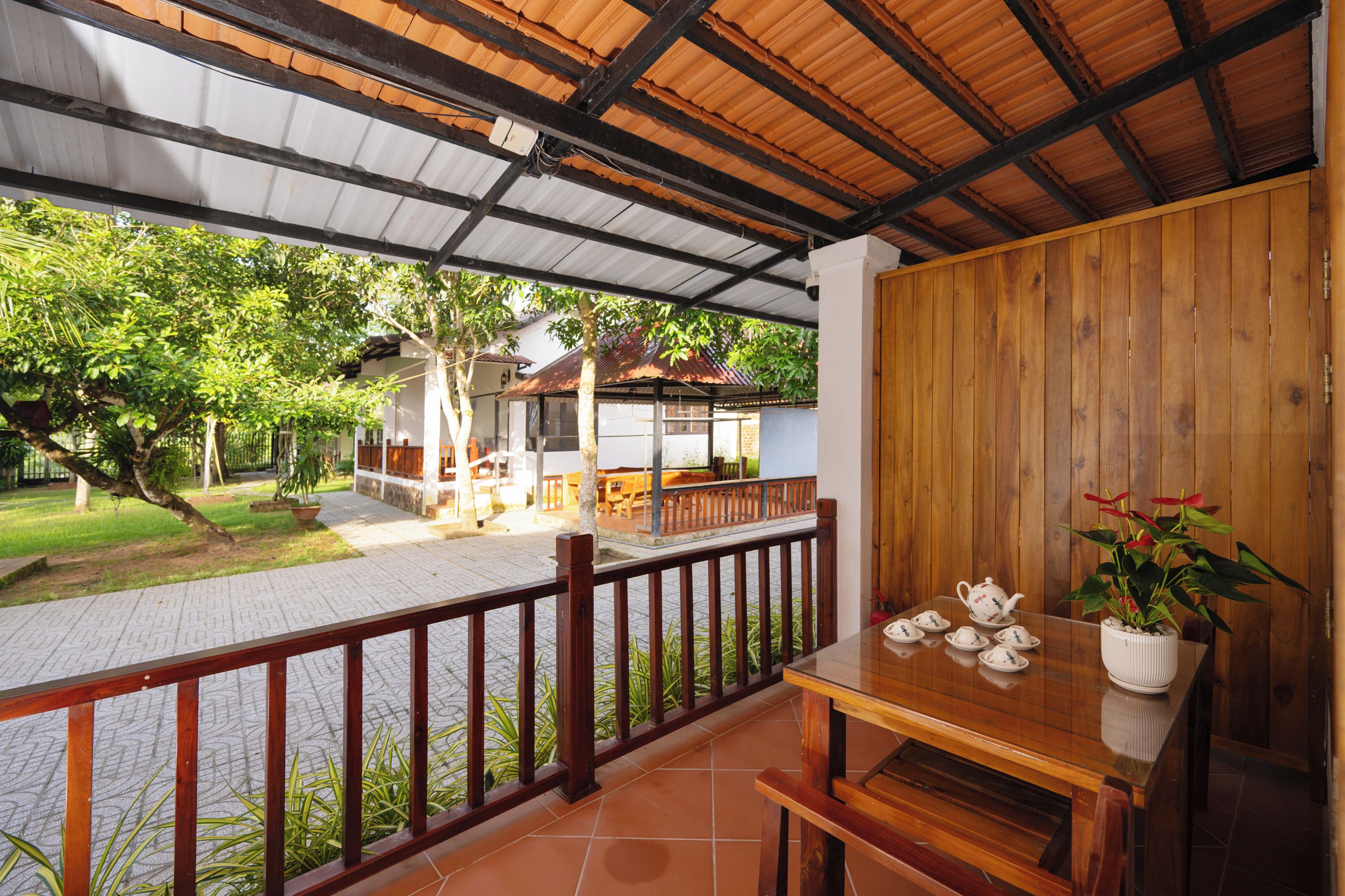 A covered outdoor area with a wooden table and chairs, a railing, and a view of a building and trees.