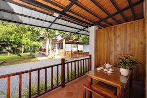 A covered outdoor area with a wooden table and chairs, a railing, and a view of a building and trees.
