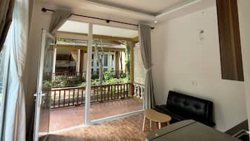 Una habitación con puerta corrediza de vidrio que da a un balcón exterior con piso de madera y vista a una casa y vegetación.