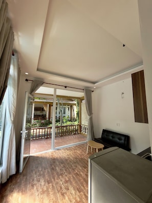 Una habitación con puerta corrediza de vidrio que da a un balcón exterior con piso de madera y vista a una casa y vegetación.