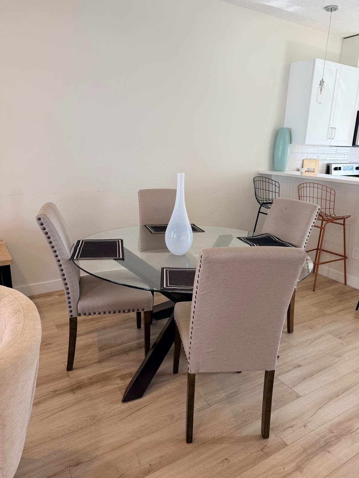 Een eetgedeelte met een glazen tafel, vier stoelen, een witte vaas en een keuken op de achtergrond.