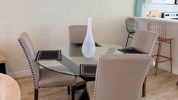 Een eetgedeelte met een glazen tafel, vier stoelen, een witte vaas en een keuken op de achtergrond.