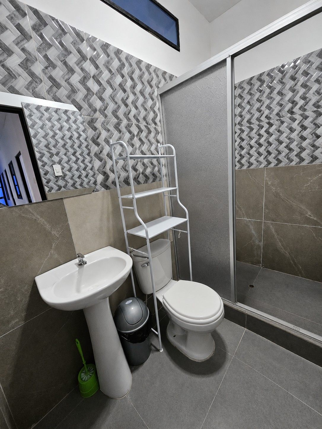 Une salle de bain avec un lavabo, des toilettes et une douche, le tout de couleur blanche. On trouve une étagère métallique à côté des toilettes et une brosse de nettoyage verte sur le sol.