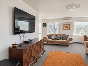 Ein Wohnzimmer mit Flachbildfernseher an der Wand, einer Couch mit gemusterten Kissen, einer Holzkommode und einem runden Teppich.