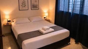 Una cama bien hecha con edredón blanco y una toalla doblada, dos lámparas de noche y un cuadro en la pared.
