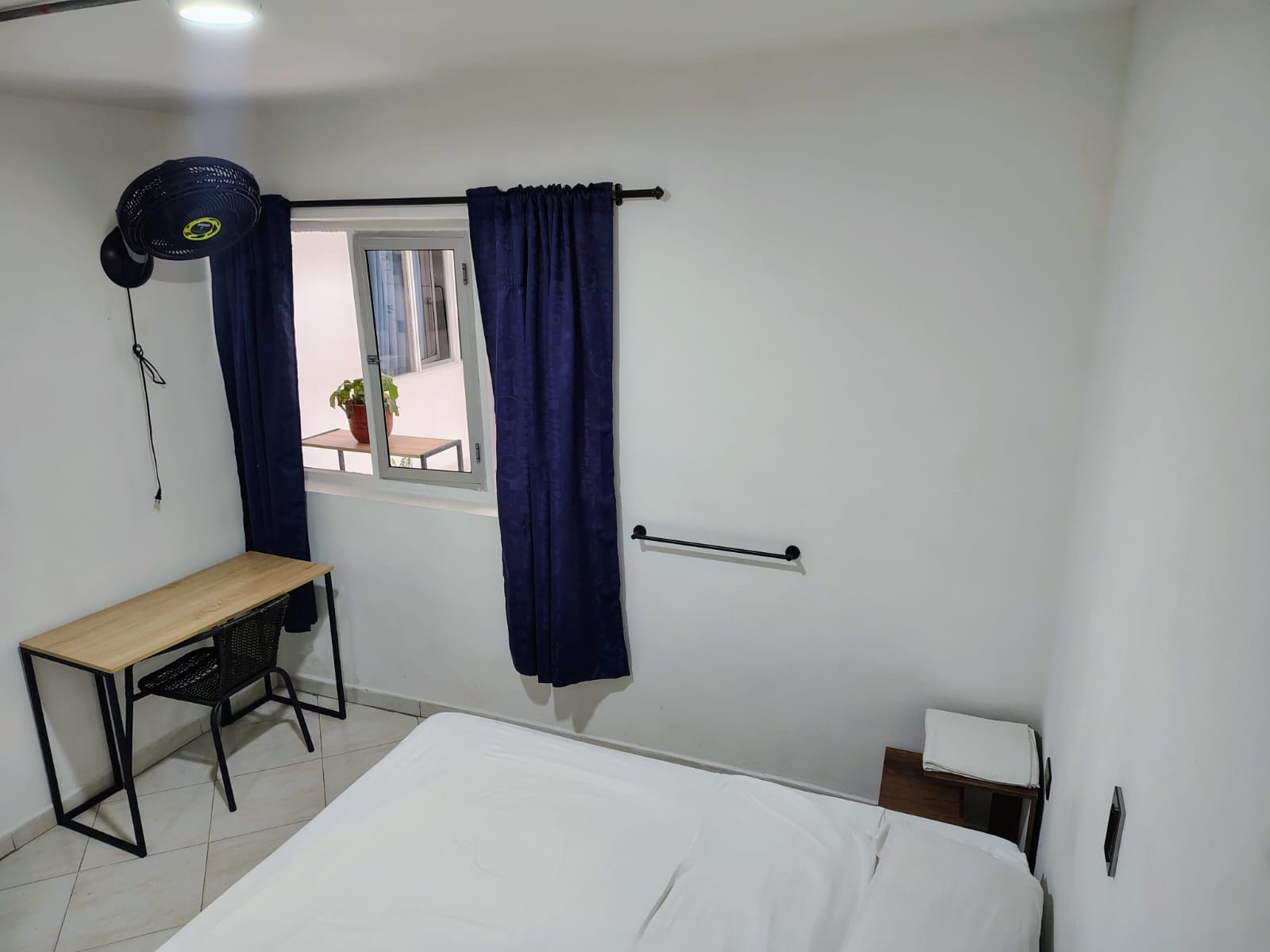 Un dormitorio con cama, escritorio, silla, ventana con cortinas y una planta en maceta afuera.