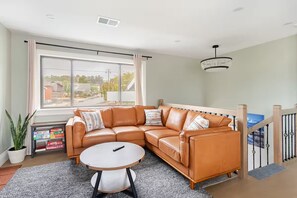A living room with a sectional sofa, a round coffee table, a potted plant, and a view through a large window. - Stylish Apartment Near Beaches, Trails, & Restaurants (El Granada)