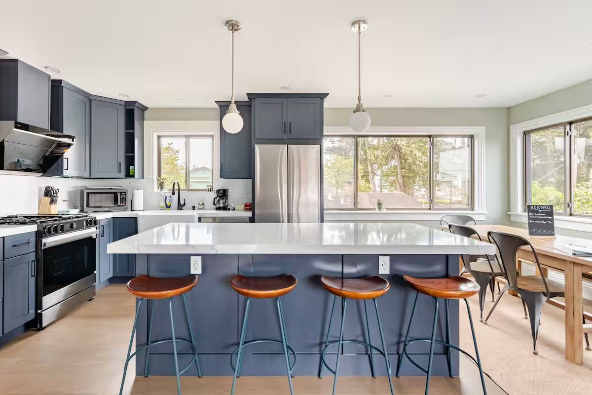 A modern kitchen with a central island, bar stools, a stainless steel refrigerator, and a large window providing natural light.