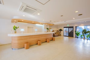 A reception area with a wooden reception desk, potted plants, and a large window. - Mango Monthly Miyazaki (Miyazaki Tachibanadorihigashi)