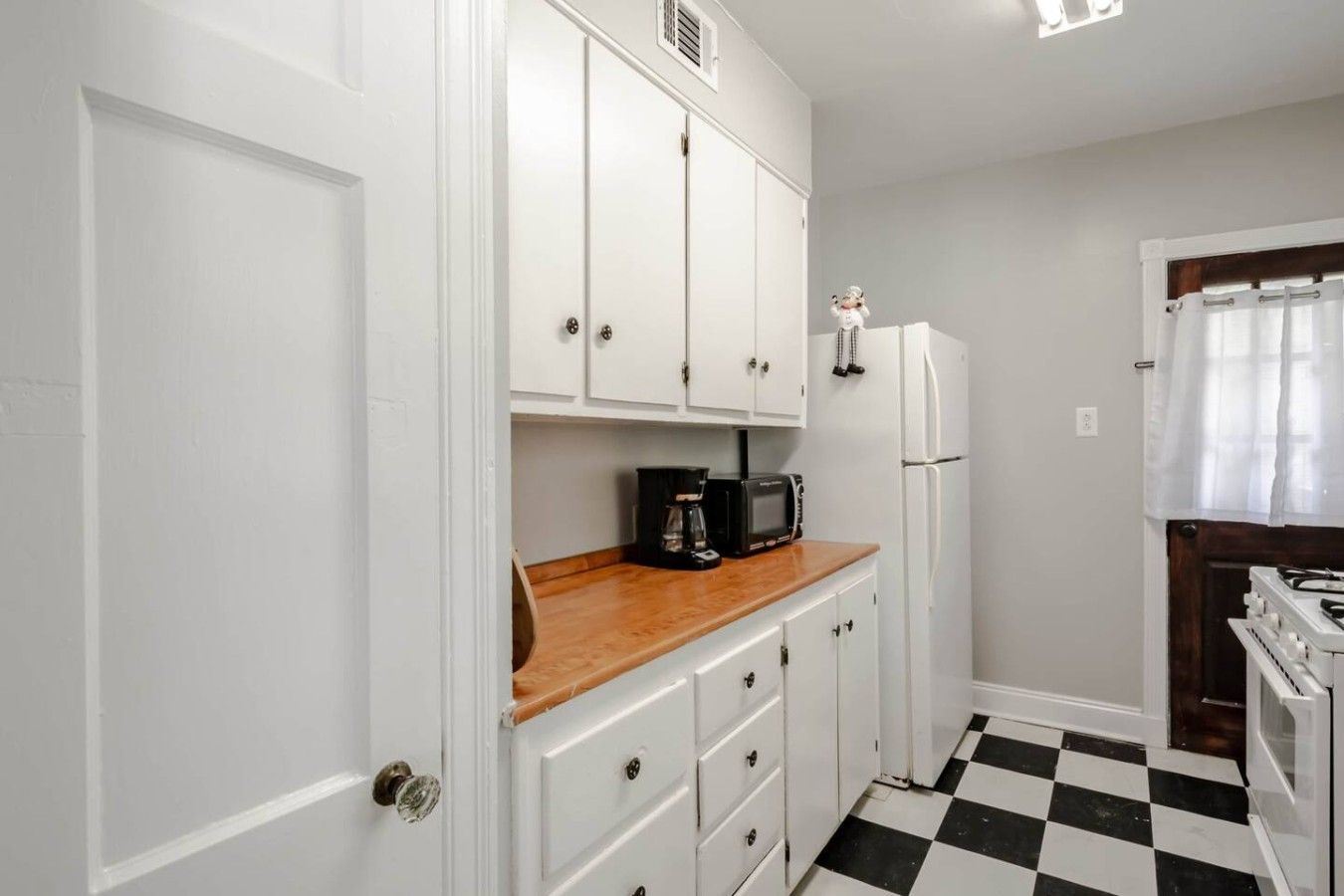Una cocina compacta con armarios blancos, encimera de madera, cafetera negra, refrigerador blanco y piso a cuadros.
