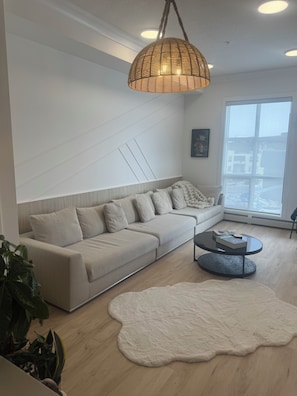 A modern living room with a sectional sofa, a round coffee table, and a large window providing a view of the city. - Luxury Elegant Condo Prime Location & Parking (Calgary)