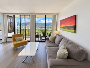 Una sala moderna con un sofá, una mesa de centro y un sillón, un cuadro en la pared y un balcón con vistas a la ciudad y a las montañas.