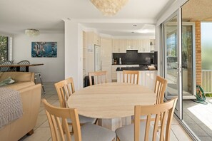 A modern kitchen with a central island, a dining area with a round table and chairs, and a sliding glass door leading to a balcony. - Shore Thing | Blue Monaro Beachfront Bliss (Burleigh Heads)
