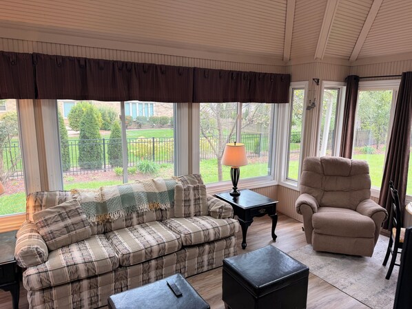 A room with a sofa, armchair, and ottoman, a lamp, and large windows with a view of a garden.